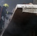 A firefighter in full protective gear is climbing a metal ladder toward the roof or upper level of a damaged concrete structure surrounded by heavy smoke. The firefighter is equipped with a self-contained breathing apparatus (SCBA) tank on their back and is carrying a fire hose. The scene depicts an active emergency response operation in a smoke-filled environment.