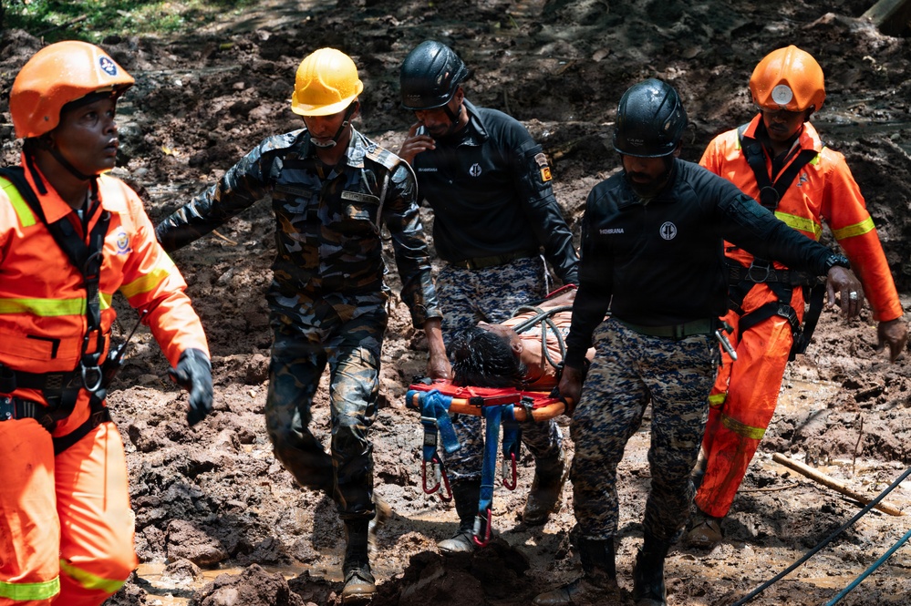 Rescue personnel are carrying an injured or unconscious person on a stretcher through muddy terrain, likely following a landslide or natural disaster. The team consists of multiple responders wearing different uniforms including military camouflage and orange rescue suits. The scene depicts an active emergency rescue operation in difficult ground conditions.