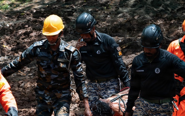 Rescue personnel are carrying an injured or unconscious person on a stretcher through muddy terrain, likely following a landslide or natural disaster. The team consists of multiple responders wearing different uniforms including military camouflage and orange rescue suits. The scene depicts an active emergency rescue operation in difficult ground conditions.