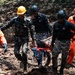 Rescue personnel are carrying an injured or unconscious person on a stretcher through muddy terrain, likely following a landslide or natural disaster. The team consists of multiple responders wearing different uniforms including military camouflage and orange rescue suits. The scene depicts an active emergency rescue operation in difficult ground conditions.