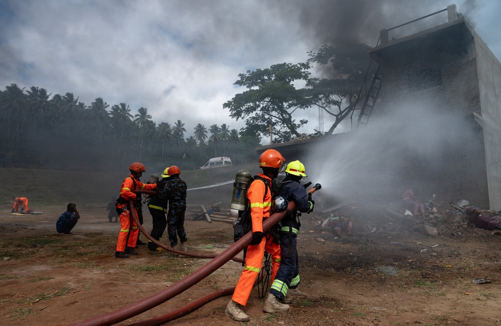 Firefighters in orange uniforms and helmets are actively battling a structural fire at what appears to be a building or warehouse. Multiple personnel are working together managing fire hoses while thick black and white smoke billows from the structure. An ambulance is visible in the background, and several civilians appear to be seated or crouching nearby.