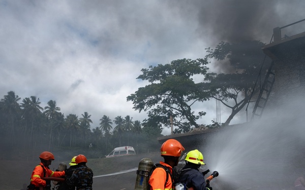 Firefighters in orange uniforms and helmets are actively battling a structural fire at what appears to be a building or warehouse. Multiple personnel are working together managing fire hoses while thick black and white smoke billows from the structure. An ambulance is visible in the background, and several civilians appear to be seated or crouching nearby.