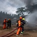 Firefighters in orange uniforms and helmets are actively battling a structural fire at what appears to be a building or warehouse. Multiple personnel are working together managing fire hoses while thick black and white smoke billows from the structure. An ambulance is visible in the background, and several civilians appear to be seated or crouching nearby.