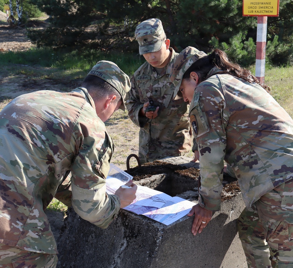 Three U.S. Army soldiers in MultiCam/OCP camouflage uniforms are conducting land navigation training at what appears to be a military installation in Poland. They are gathered around a concrete structure examining maps and using a compass. A yellow warning sign in Polish is visible in the background.