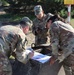 Three U.S. Army soldiers in MultiCam/OCP camouflage uniforms are conducting land navigation training at what appears to be a military installation in Poland. They are gathered around a concrete structure examining maps and using a compass. A yellow warning sign in Polish is visible in the background.