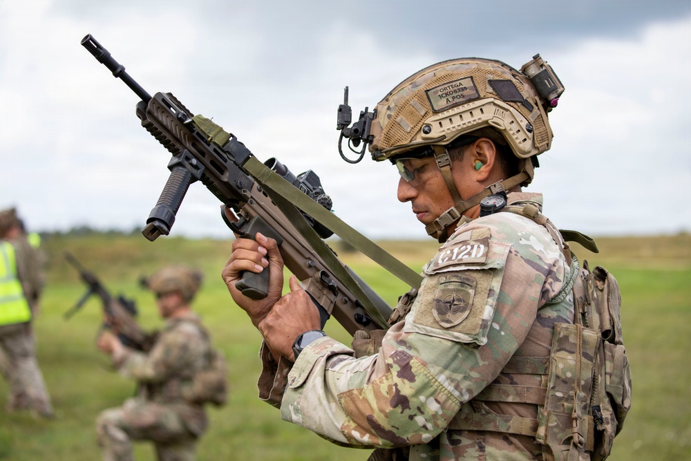 A soldier in full tactical gear examines a bullpup-style assault rifle in an open field environment. The soldier wears a NATO patch and multicam camouflage uniform with a tactical helmet equipped with communications gear. Additional personnel are visible in the background, suggesting a training exercise or field demonstration.