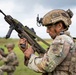 A soldier in full tactical gear examines a bullpup-style assault rifle in an open field environment. The soldier wears a NATO patch and multicam camouflage uniform with a tactical helmet equipped with communications gear. Additional personnel are visible in the background, suggesting a training exercise or field demonstration.