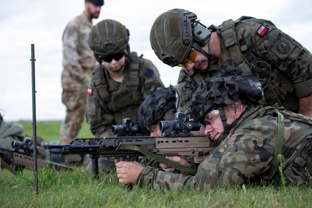 Military personnel in camouflage uniforms are conducting a shooting exercise on a grass range. A prone shooter is aiming a rifle with an optical scope while a standing soldier observes and coaches from above. Polish flag patches are visible on multiple uniforms, indicating Polish Armed Forces participation, with at least one soldier in a different camouflage pattern suggesting a multinational exercise.
