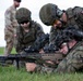 Military personnel in camouflage uniforms are conducting a shooting exercise on a grass range. A prone shooter is aiming a rifle with an optical scope while a standing soldier observes and coaches from above. Polish flag patches are visible on multiple uniforms, indicating Polish Armed Forces participation, with at least one soldier in a different camouflage pattern suggesting a multinational exercise.