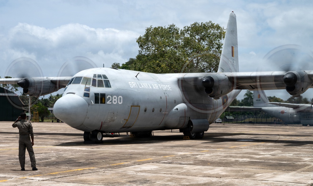 A Sri Lanka Air Force C-130 Hercules transport aircraft (serial number 280) is parked on a concrete apron at a military airbase. A uniformed personnel member stands in front of the aircraft, appearing to photograph or observe it. The propellers are spinning, indicating the engines are running or have recently been started.