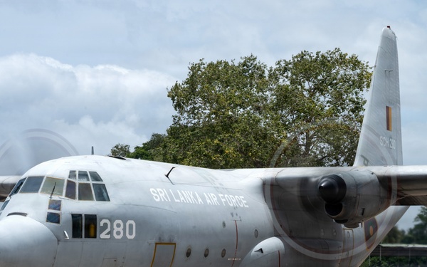 A Sri Lanka Air Force C-130 Hercules transport aircraft (serial number 280) is parked on a concrete apron at a military airbase. A uniformed personnel member stands in front of the aircraft, appearing to photograph or observe it. The propellers are spinning, indicating the engines are running or have recently been started.