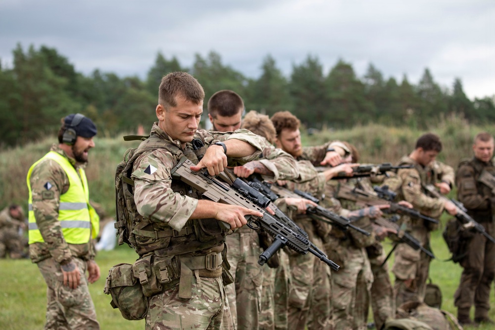 A group of military personnel in camouflage uniforms are conducting a weapons handling or drill exercise in an outdoor field setting. Soldiers are lined up in a row, each holding and inspecting their rifles in a synchronized manner. A safety observer wearing a high-visibility yellow vest is present on the left side of the formation.