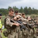 A group of military personnel in camouflage uniforms are conducting a weapons handling or drill exercise in an outdoor field setting. Soldiers are lined up in a row, each holding and inspecting their rifles in a synchronized manner. A safety observer wearing a high-visibility yellow vest is present on the left side of the formation.