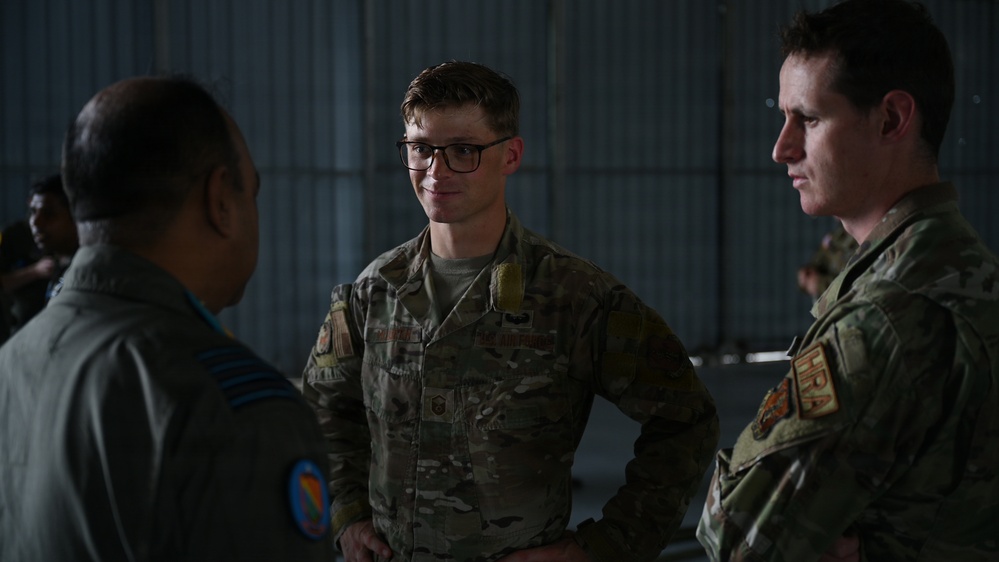 Three military personnel are engaged in conversation inside what appears to be a hangar or military facility. The central figure wears US Air Force OCP (Operational Camouflage Pattern) uniform with visible rank insignia and name tape, while the figure on the left wears a different uniform with a squadron patch, suggesting a multinational or joint-service interaction. The setting and attire suggest a professional military briefing or exchange.