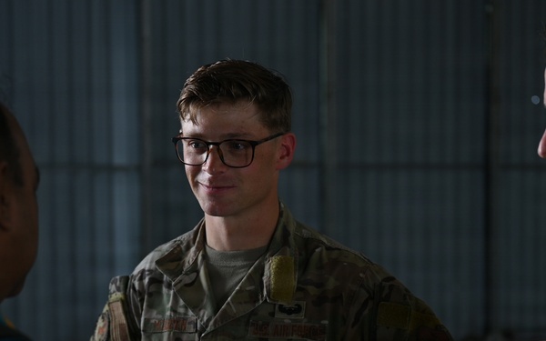 Three military personnel are engaged in conversation inside what appears to be a hangar or military facility. The central figure wears US Air Force OCP (Operational Camouflage Pattern) uniform with visible rank insignia and name tape, while the figure on the left wears a different uniform with a squadron patch, suggesting a multinational or joint-service interaction. The setting and attire suggest a professional military briefing or exchange.