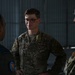 Three military personnel are engaged in conversation inside what appears to be a hangar or military facility. The central figure wears US Air Force OCP (Operational Camouflage Pattern) uniform with visible rank insignia and name tape, while the figure on the left wears a different uniform with a squadron patch, suggesting a multinational or joint-service interaction. The setting and attire suggest a professional military briefing or exchange.