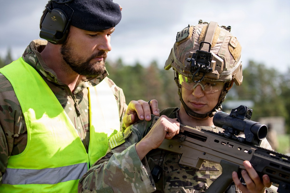 Two military personnel are engaged in what appears to be a weapons instruction or inspection session in an outdoor training environment. One individual wearing a high-visibility yellow vest, black beret, and hearing protection appears to be an instructor or safety officer, while the other is a fully equipped soldier wearing a tactical helmet with night vision mount, ballistic eyewear, and holding a bullpup-style assault rifle. The setting suggests a military training exercise or range activity.