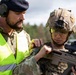 Two military personnel are engaged in what appears to be a weapons instruction or inspection session in an outdoor training environment. One individual wearing a high-visibility yellow vest, black beret, and hearing protection appears to be an instructor or safety officer, while the other is a fully equipped soldier wearing a tactical helmet with night vision mount, ballistic eyewear, and holding a bullpup-style assault rifle. The setting suggests a military training exercise or range activity.