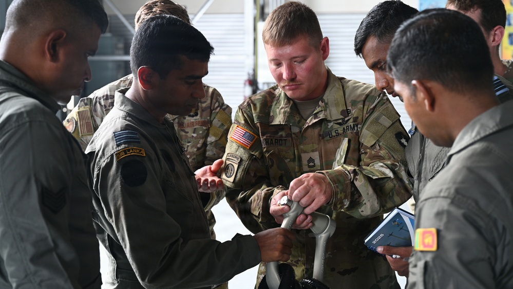 A U.S. Army soldier wearing 82nd Airborne Division patches is demonstrating or explaining a piece of equipment to Sri Lanka Air Force personnel. The interaction appears to be part of a joint military training or exchange program. Multiple uniformed personnel from both nations are gathered closely, examining the item together.