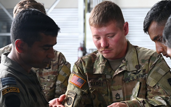 A U.S. Army soldier wearing 82nd Airborne Division patches is demonstrating or explaining a piece of equipment to Sri Lanka Air Force personnel. The interaction appears to be part of a joint military training or exchange program. Multiple uniformed personnel from both nations are gathered closely, examining the item together.