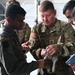A U.S. Army soldier wearing 82nd Airborne Division patches is demonstrating or explaining a piece of equipment to Sri Lanka Air Force personnel. The interaction appears to be part of a joint military training or exchange program. Multiple uniformed personnel from both nations are gathered closely, examining the item together.