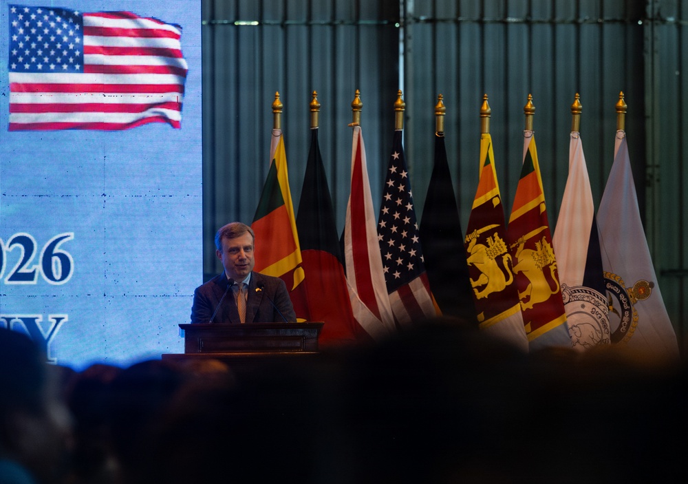 A man in a suit is speaking at a podium during what appears to be a formal diplomatic or official event. Multiple national flags are displayed behind him, including the United States and Sri Lanka flags, alongside a large projected US flag on a screen to the left. Partial text visible on the screen includes '026' and 'Y', suggesting a year reference possibly related to 2026.