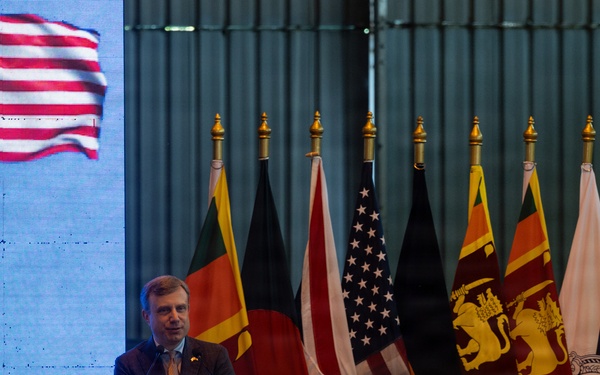 A man in a suit is speaking at a podium during what appears to be a formal diplomatic or official event. Multiple national flags are displayed behind him, including the United States and Sri Lanka flags, alongside a large projected US flag on a screen to the left. Partial text visible on the screen includes '026' and 'Y', suggesting a year reference possibly related to 2026.