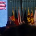 A man in a suit is speaking at a podium during what appears to be a formal diplomatic or official event. Multiple national flags are displayed behind him, including the United States and Sri Lanka flags, alongside a large projected US flag on a screen to the left. Partial text visible on the screen includes '026' and 'Y', suggesting a year reference possibly related to 2026.