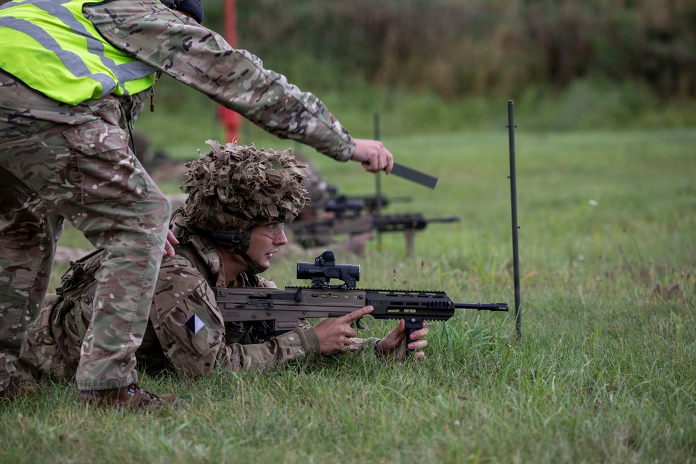 A military training exercise on a grass range where a prone soldier in camouflage uniform and ghillie-style helmet cover aims a bullpup assault rifle, likely an SA80/L85A3. A standing instructor wearing a high-visibility yellow safety vest over MultiCam uniform provides guidance, holding what appears to be a range flag or marker. Additional soldiers with weapons are visible in the background, indicating a supervised live-fire or marksmanship training session.