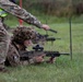 A military training exercise on a grass range where a prone soldier in camouflage uniform and ghillie-style helmet cover aims a bullpup assault rifle, likely an SA80/L85A3. A standing instructor wearing a high-visibility yellow safety vest over MultiCam uniform provides guidance, holding what appears to be a range flag or marker. Additional soldiers with weapons are visible in the background, indicating a supervised live-fire or marksmanship training session.