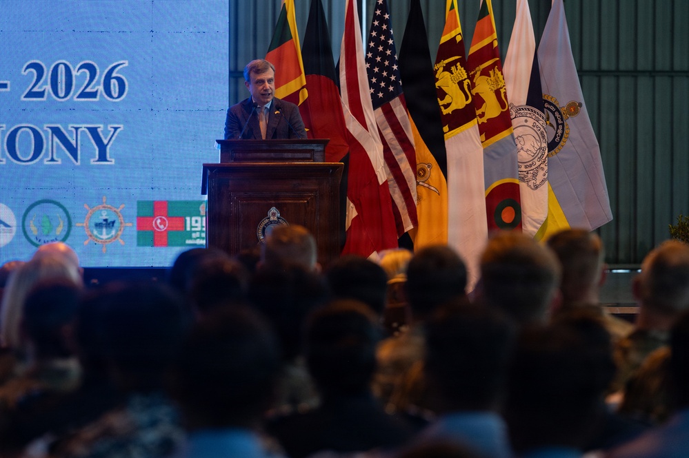 A male speaker addresses a large audience from a wooden podium during a formal ceremony, with a backdrop displaying '2026' and 'ONY' (likely 'CEREMONY'). Multiple national and organizational flags are displayed behind him, including those of the United States, United Kingdom, Sri Lanka, and Bangladesh. The event appears to be a multinational official or military ceremony.