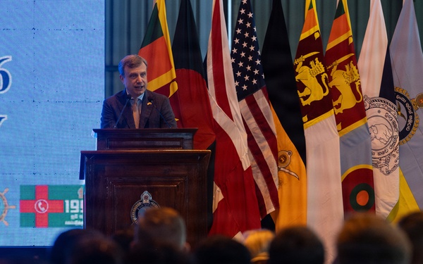 A male speaker addresses a large audience from a wooden podium during a formal ceremony, with a backdrop displaying '2026' and 'ONY' (likely 'CEREMONY'). Multiple national and organizational flags are displayed behind him, including those of the United States, United Kingdom, Sri Lanka, and Bangladesh. The event appears to be a multinational official or military ceremony.