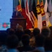 A male speaker addresses a large audience from a wooden podium during a formal ceremony, with a backdrop displaying '2026' and 'ONY' (likely 'CEREMONY'). Multiple national and organizational flags are displayed behind him, including those of the United States, United Kingdom, Sri Lanka, and Bangladesh. The event appears to be a multinational official or military ceremony.