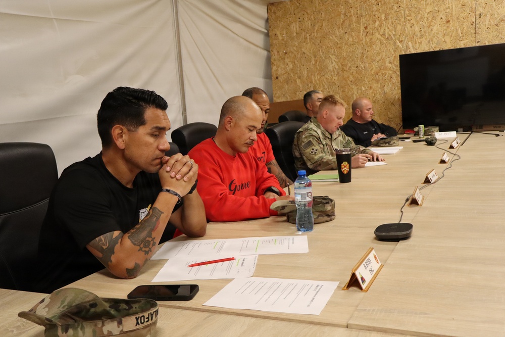 A group of approximately six individuals are seated along one side of a long conference table in what appears to be a military forward operating base or temporary facility. The group includes both uniformed military personnel and civilians engaged in what appears to be a briefing or planning meeting. The setting features plywood walls, a television monitor, name placards on the table, and various documents.