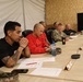 A group of approximately six individuals are seated along one side of a long conference table in what appears to be a military forward operating base or temporary facility. The group includes both uniformed military personnel and civilians engaged in what appears to be a briefing or planning meeting. The setting features plywood walls, a television monitor, name placards on the table, and various documents.