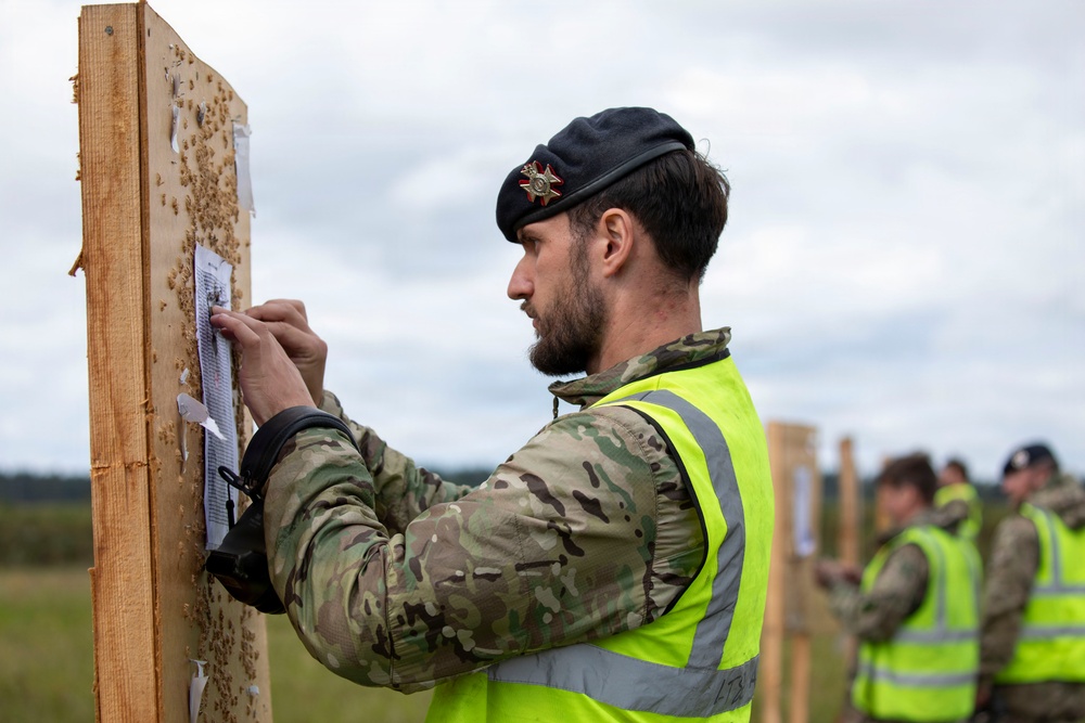 A military personnel member wearing camouflage uniform, a black beret with a badge, and a high-visibility yellow safety vest is attaching or reviewing a paper document on a wooden board at what appears to be a shooting range. In the background, other similarly dressed individuals in high-visibility vests are performing the same task at adjacent wooden boards. The setting appears to be an outdoor military training or competition range under overcast skies.