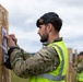 A military personnel member wearing camouflage uniform, a black beret with a badge, and a high-visibility yellow safety vest is attaching or reviewing a paper document on a wooden board at what appears to be a shooting range. In the background, other similarly dressed individuals in high-visibility vests are performing the same task at adjacent wooden boards. The setting appears to be an outdoor military training or competition range under overcast skies.