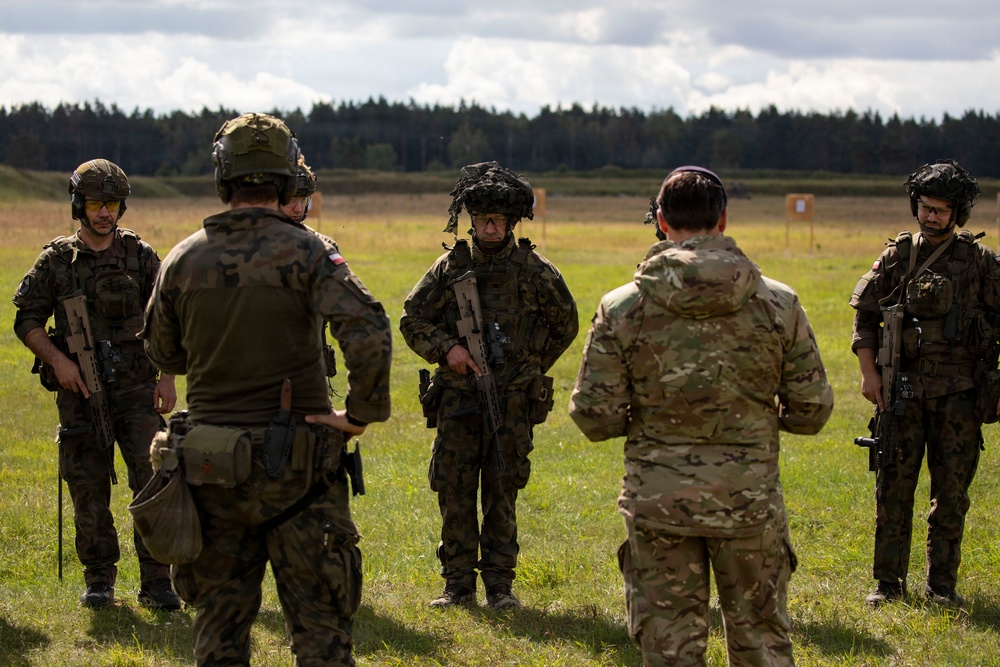 A group of military personnel in full combat gear are gathered in an open field, appearing to receive a briefing or debrief from one soldier facing away from the camera. At least two distinct camouflage patterns are visible, suggesting a multinational or joint training exercise. A shooting range target is visible in the background, indicating a live-fire or marksmanship training environment.