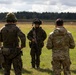 A group of military personnel in full combat gear are gathered in an open field, appearing to receive a briefing or debrief from one soldier facing away from the camera. At least two distinct camouflage patterns are visible, suggesting a multinational or joint training exercise. A shooting range target is visible in the background, indicating a live-fire or marksmanship training environment.