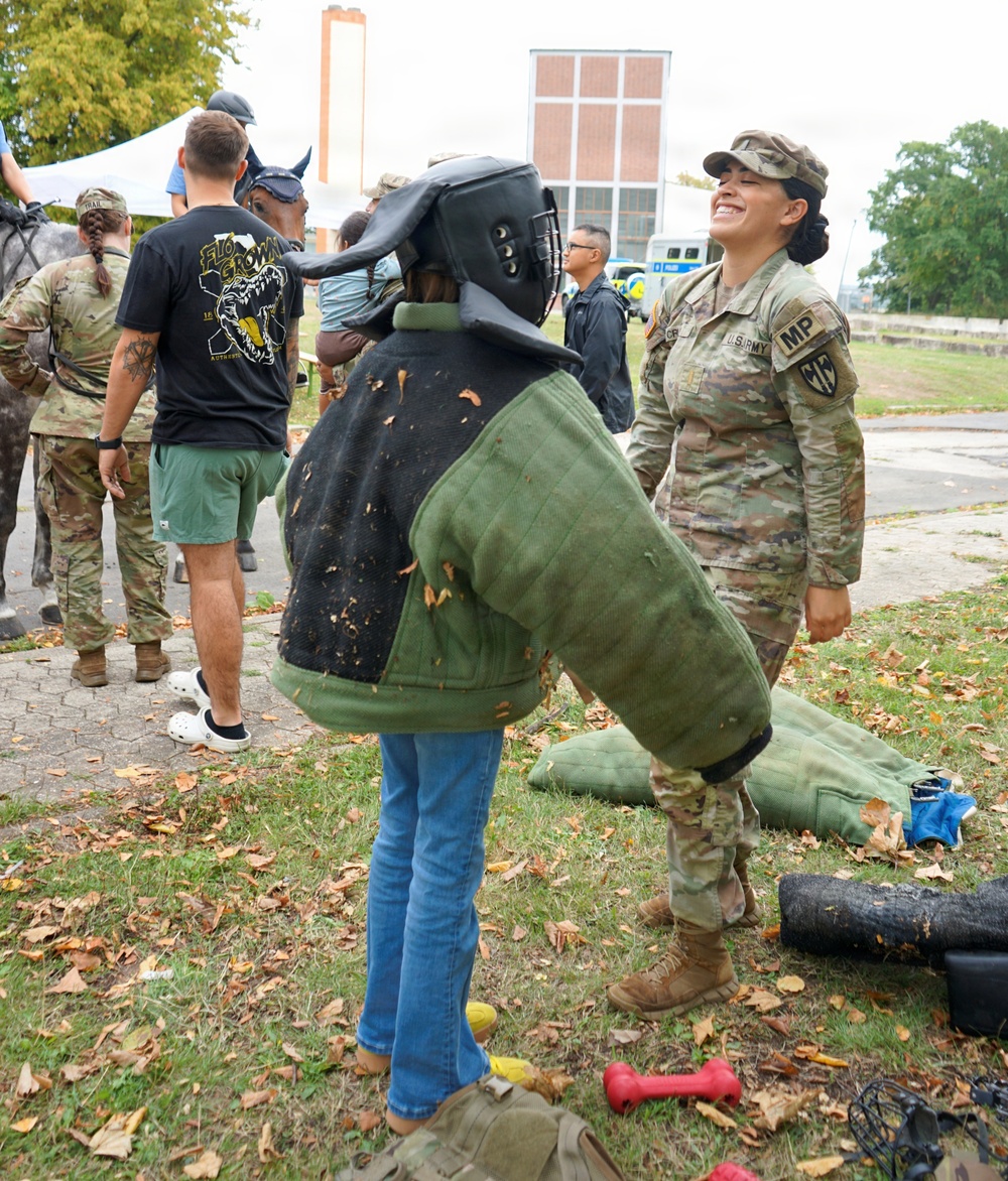 A U.S. Army Military Police (MP) soldier smiles while a child tries on a bite suit (K9 training protective gear) including a padded jacket and helmet with face guard. The scene appears to be an open house or community outreach event at a military installation, with other soldiers, civilians, and a horse visible in the background. Fallen leaves on the ground suggest an autumn setting.