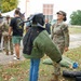 A U.S. Army Military Police (MP) soldier smiles while a child tries on a bite suit (K9 training protective gear) including a padded jacket and helmet with face guard. The scene appears to be an open house or community outreach event at a military installation, with other soldiers, civilians, and a horse visible in the background. Fallen leaves on the ground suggest an autumn setting.