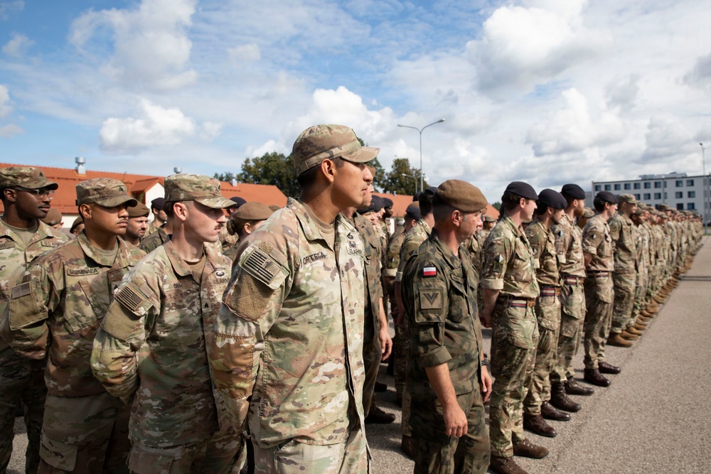 A formation of multinational military personnel standing at attention during what appears to be a formal military ceremony or parade. Soldiers from at least two identifiable nations are present, including US Army personnel and what appears to be Polish military forces, identifiable by a Polish flag patch. The formation is taking place outdoors on a paved surface near military or institutional buildings.