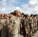 A formation of multinational military personnel standing at attention during what appears to be a formal military ceremony or parade. Soldiers from at least two identifiable nations are present, including US Army personnel and what appears to be Polish military forces, identifiable by a Polish flag patch. The formation is taking place outdoors on a paved surface near military or institutional buildings.