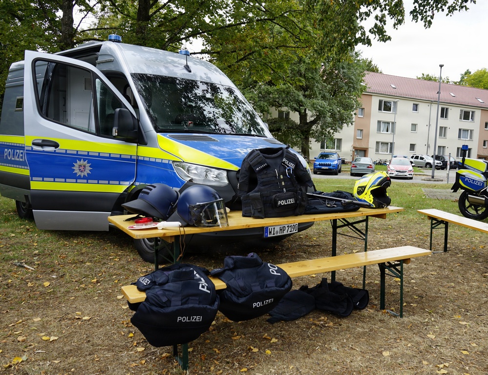 A German police (Polizei) equipment display is set up outdoors on wooden picnic tables in front of a blue and yellow police van. Various tactical gear items are laid out for what appears to be a public demonstration or open day event. A police motorcycle is also visible in the background on the right side.