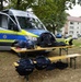 A German police (Polizei) equipment display is set up outdoors on wooden picnic tables in front of a blue and yellow police van. Various tactical gear items are laid out for what appears to be a public demonstration or open day event. A police motorcycle is also visible in the background on the right side.