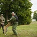 A military working dog (MWD) training exercise is taking place outdoors on a grassy field. A U.S. Army soldier in MultiCam/OCP uniform and patrol cap is handling a dog on a leash while the dog bites or engages a decoy wearing a padded green bite suit. The setting appears to be a training area with trees in the background under an overcast sky.