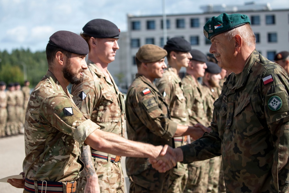A military ceremony appears to be taking place outdoors, with a senior officer wearing a green beret and Polish flag patch shaking hands with soldiers in multicam/MTP camouflage uniforms. The scene suggests a multinational military event, likely involving Polish and British armed forces personnel standing in formation. The setting appears to be a military installation or training ground.