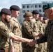 A military ceremony appears to be taking place outdoors, with a senior officer wearing a green beret and Polish flag patch shaking hands with soldiers in multicam/MTP camouflage uniforms. The scene suggests a multinational military event, likely involving Polish and British armed forces personnel standing in formation. The setting appears to be a military installation or training ground.