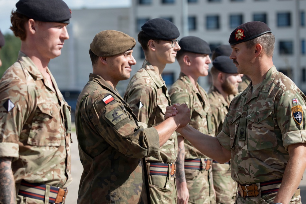 A military ceremony or inspection is taking place outdoors, with a senior British officer shaking hands with a Polish soldier standing in a line formation. The soldiers wear a mix of British MTP (Multi-Terrain Pattern) and Polish camouflage uniforms, with visible national flag patches and NATO insignia. The setting appears to be a military base or parade ground with a building visible in the background.