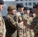 A military ceremony or inspection is taking place outdoors, with a senior British officer shaking hands with a Polish soldier standing in a line formation. The soldiers wear a mix of British MTP (Multi-Terrain Pattern) and Polish camouflage uniforms, with visible national flag patches and NATO insignia. The setting appears to be a military base or parade ground with a building visible in the background.